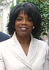 A woman with short wavy hair smiles, and is wearing a white jacket.