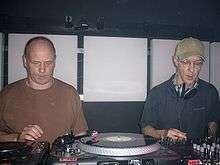 Two men perform music using various mixing boards and turntables.