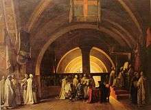 Painting of dozens of men in white robes in a domed chapel. A young man with short black hair and dark blue robe is kneeling on a stool in the center, and pointing at something in a large open book which is being held by one of the white-robed men.