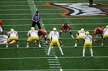 Two football teams line up against each other on a green football field. The foreground, offensive team has yellow helmets, white jerseys, yellow pants, white socks, and yellow and black shoes. The opposing, defensive team has black helmets, burnt orange jerseys, white pants and socks, and orange and black shoes.