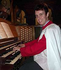 Colour photograph of smiling organist in red and white vestments playing the St James' organ. The sheet music, keyboard and some of the organ stops are visible.