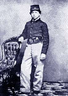 A white adolescent boy standing with his right arm resting on the back of a chair. He is wearing a forage cap, a dark military jacket, and light-colored pants.