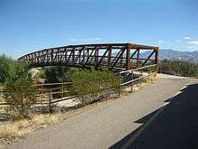 Photo shows a rust-colored truss footbridge with mountains in the right background.