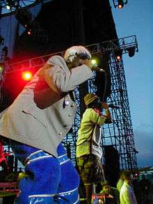 Two African-American men rap into microphones whilst on stage. One wears a blonde wig, a grey jacket and blue trousers, and the other wears a green checkered hat, a white shirt and khaki shorts.
