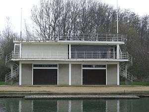 A two-storey building on a river bank. There are railings at balcony and roof level, two flag poles (one on the far left of the roof, one on the far right), external spiral staircases leading to roof level (one on the far left of the building, the other on the far right) and two large doors at ground level at the front.