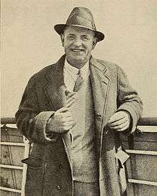 middle-aged man in overcoat and trilby hat smiling cheerfully towards the camera