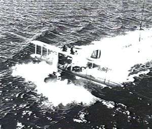 Single-engined military biplane on floats landing in ocean and trailing heavy wake, three-quarters overhead