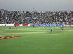 An image of a cricket field with the spectators in background. Players in blue outfit can be seen fielding, and a player in a red-blue outfit can be seen batting.