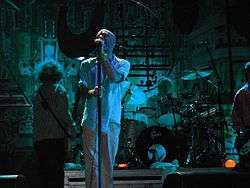 A blue-tinted photograph of musicians in front of an industrial background. From left to right: a long-haired male stands with his back to the camera playing bass guitar, a middle-aged Caucasian male sings into a microphone, a middle-aged Caucasian male plays behind a black-and-silver drum set on a riser, and a guitar player is mostly cropped from the extreme left of the photo.