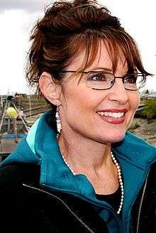 A woman wearing glasses, earrings, red lipstick, a necklace of white beads, and a black jacket on top of a blue hoodie