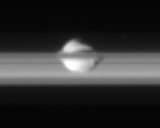 A bright fuzzy band (rings of Saturn) is running from the left to right. In the center a bright irregularity shaped body is superimposed on its upper edge. A narrow grayish band, which is a part of the main band, partially covers the body.