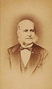 A photograph showing the head and shoulders of an older, balding man with long sideburns dressed in a dark suit, vest and bow tie