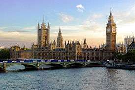 Neo-gothic Westminister palace and Big Ben clock tower stand above a river and bridge.