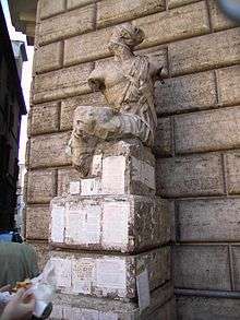 A damaged statue is on a pedestal in front of a stone wall; the pedestal has a number of pieces of paper with writing on them glued to it