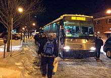 A line of people, seen from behind, boarding two buses. The nearer one has "MNR service" on its front display. It is dark, with orangish-colored streetlights illuminating snow