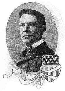 Head of a white man wearing a dark suit and bow tie. To the lower right of the oval-shaped portrait is an illustration of a shield covered with stars and stripes, like the American flag, and a ribbon tied around the shield.