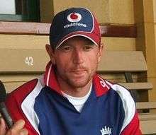 An English cricketer in a red, white and blue shirt and cap, with his tongue between his lips.