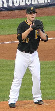 A man in a black baseball jersey, white pants, and black baseball cap wearing a black baseball glove on his right hand preparing to pitch from a baseball mound.