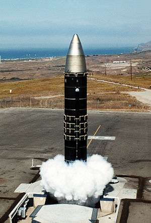 Huge missile with body painted in black rising out of silo during a launch, producing clouds of gas at the silo's opening. In the distant is a coastline.