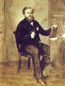 Photograph of a man with a full beard and dressed in a dark frock coat who is seated at a table holding a book with bookshelves in the background