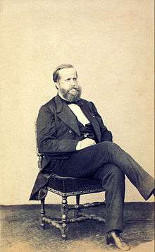 Photograph of a seated, bearded man dressed in a dark suit and vest