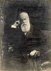 Photographic portrait of a man with a white beard seated in an armchair and holding a small book in his left hand while supporting his head with his right hand