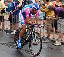 A road racing cyclist in a pink and purple skinsuit with white trim and an aerodynamic helmet. Numerous spectators stand at the roadside behind a metal barricade.