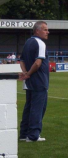 A middle-aged man with greying hair, wearing a dark blue T-shirt and matching jogging bottoms, standing near the touchline of a sports pitch.  Some spectators are visible in the background.
