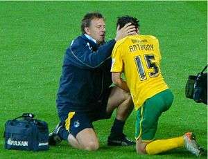 A man with short, dark hair wearing a navy blue jacket, navy blue shorts with a yellow stripe, and dark coloured shoes and socks kneeling on one knee. His hands are on the sides of the head of a second man who has his back to the camera. He is also kneeling on one knee, has dark hair and is wearing a yellow football shirt with the name 'Anthony' written across the shoulders and a large number 15 in the centre of the back. He is wearing green shorts, long yellow socks, and light-coloured football boots. Both men are on a grass surface.