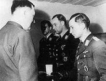 Three men all wearing military uniforms and decorations standing in row. The man on the right is taking a box from another man whose back is facing the camera.