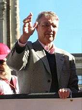 A white-haired man in a tweed jacket, dark sweater, and a pink dress shirt waves from atop a truck.