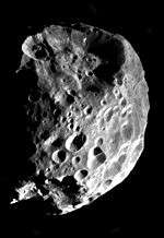 An approximately spherical heavily cratered body is illuminated from the bottom-right. The terminator runs near the left and top limbs. There is huge crater at the top, which affects the shape, and another slightly smaller at the bottom.