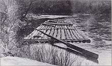 A raft made of long logs lashed together with a large oar for steering is tied to the bank of a stream