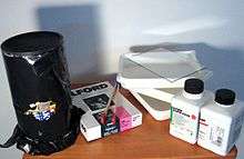 A pinhole camera made from an oatmeal container, wrapped in opaque plastic to prevent light leaks; a box of photographic paper; tongs and dishes for developing film; bottles of film developing chemicals