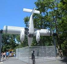 A Seneca pivoting around its wing spar and pointing vertically downwards, installed around 4 meters above a pavement in a park, with people walking underneath