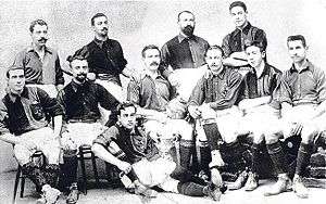 Sepia picture. 11 men pose for a squad picture, half of them bearded. Some sit on chairs, some stand and one is lying on the floor