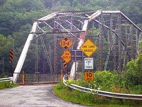 Ground-level view of a road leading up to a Pratt truss bridge spanning a river