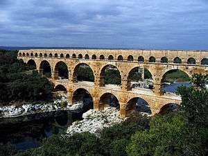A stone aqueduct consisting of three levels with many arches crosses a river.