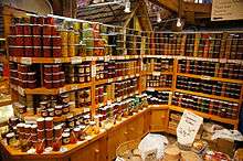 In the lower portion of the photograph is a light-coloured wooden cabinet with closed doors, above which are three levels of shelving fully stacked with glass jars containing preserves, jams, and pickled foods. Labels are affixed to the front of each wooden shelf below the stacked jars. At the top of the photograph are some of the timber support beams of the structure.