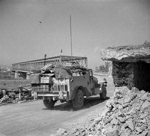Overloaded vehicle on a road heading towards a box girder bridge. A destroyed pill box is on the right