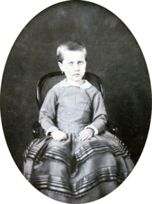 Photograph of a young Isabel with hair gathered at the back and wearing earrings seated on a small, cane back armchair