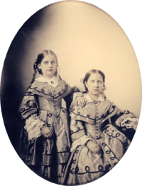 An oval, framed photographic portrait of two young girls dressed in elaborate Victorian-era gowns