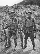 Black and white photo of two soldiers standing side by side in field uniform, each is wearing a steel helmet on their heads and load carriage equipment on their chests. Both are holding rifles in the right hands, with the weapon held vertically and the butt resting on the ground. In the background is an earthen mound.