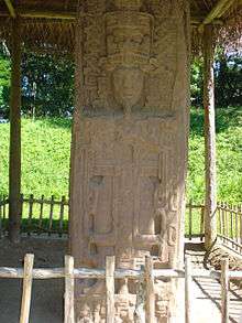 A stela with the prominent sculpture of a king surrounded by elaborate decoration. The monument is covered by a thatched roof supported on wooden poles.