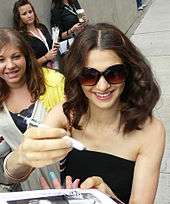 A dark-haired woman signing autographs for fans. She is wearing a black blouse and shades. Behind her there is a fan.