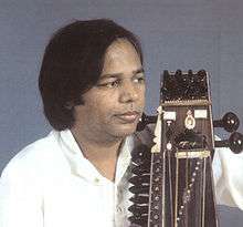 A middle-aged man wears a shirt and looks to the side with a bowed instrument held close to his body.