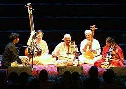 A mixed-gender group of five people sits on a platform, two playing long-necked lutes, another two playing bowed instruments, and one resting his hands next to drums.
