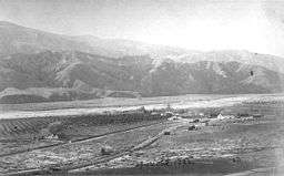 Historic photograph of Ranco Camulos from an elevated angle. The ranch spreads out on the valley bottom on the banks of a river, with mountains rising behind.