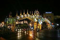 An elaborate double archway above a road, with pictures of King Bhumibol Adulyadej; trees decorated with lights