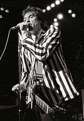 A man stands onstage with a guitar strapped across his chest. It hangs limp and unused, as he is focusing on singing into a microphone directly in front of him, which he grasps with his left hand. He wears a black-and-white, vertically striped suit.